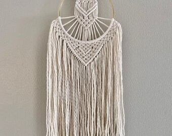 May include: A white macrame wall hanging with a gold metal hoop. The macrame has a geometric pattern and long fringe.