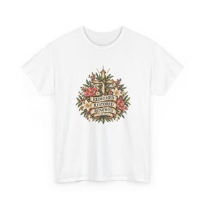 Könnte beinhalten: Ein weißes T-Shirt mit einer zentralen Grafik eines Kreuzes, das von bunten Blumen und Grünpflanzen umgeben ist. Ein beiges Banner in der Mitte zeigt den Text "REDEEMED RESTORED RENEWED" mit "BY HIS GRACE" darunter gedruckt.