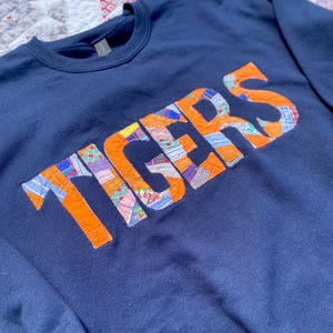 May include: Navy blue sweatshirt with the word "TIGERS" appliqued in orange fabric. The letters are filled with a patchwork design of various patterned fabrics. The sweatshirt is displayed on a patterned quilt.