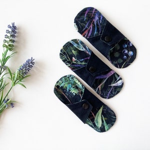 May include: Three reusable cloth menstrual pads with a dark blue background and a floral print in shades of purple, green, and white.