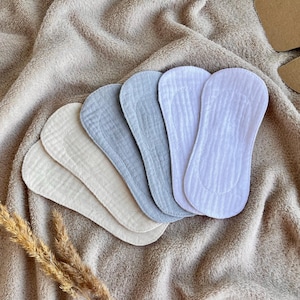 Può includere: Una collezione di assorbenti mestruali in tessuto riutilizzabili in varie tonalità di bianco, beige e blu. Gli assorbenti sono disposti su un tessuto beige morbido e strutturato, con un rametto di grano secco in primo piano. Una scatola di cartone è visibile sullo sfondo.