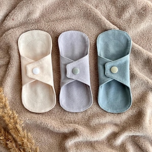 May include: Three reusable cloth menstrual pads in cream, lavender, and blue-green, each with a button closure. The pads are arranged on a textured, tan fabric background, with dried wheat stalks in the lower left corner.