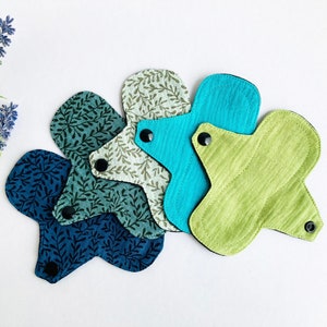 May include: Four reusable cloth menstrual pads in various shades of green and blue with a leafy pattern. The pads are arranged in a stack with the green pad on top.