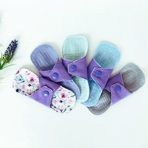 May include: Six reusable cloth menstrual pads in various shades of purple and blue with floral patterns. The pads are folded and arranged in a fan shape.