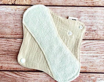 Könnte beinhalten: Eine beige wiederverwendbare Stoff-Menstruationseinlage mit einer weißen saugfähigen Schicht. Die Einlage hat zwei weiße Druckknöpfe an der Seite.