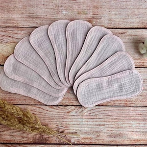 May include: A set of ten reusable cloth pads in a light pink color. The pads are arranged in a semi-circle on a wooden surface.