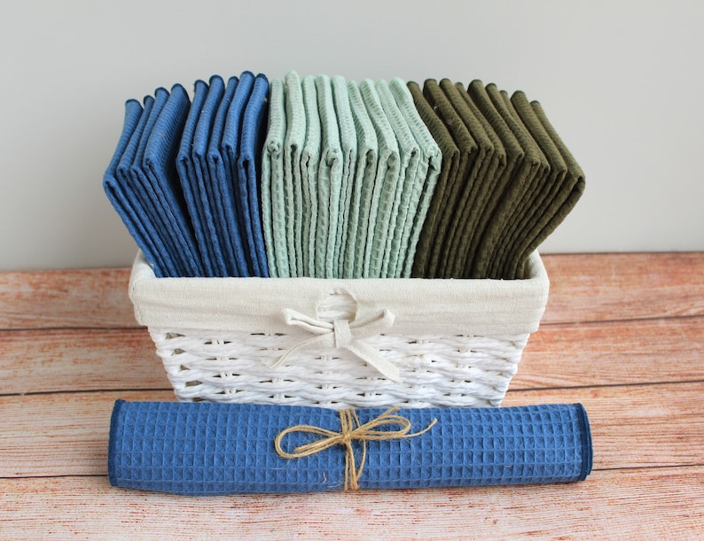 May include: A white wicker basket filled with stacks of blue, green, and olive green waffle weave kitchen towels. A rolled up blue waffle weave kitchen towel sits in front of the basket.