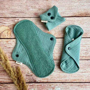 May include: Three reusable cloth menstrual pads in a teal green color. The pads are folded and laid out on a wooden surface. The pads have snaps for closure.