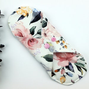 May include: A white reusable cloth menstrual pad with a floral print featuring pink, yellow, and blue watercolour flowers. The pad has two pink snaps.