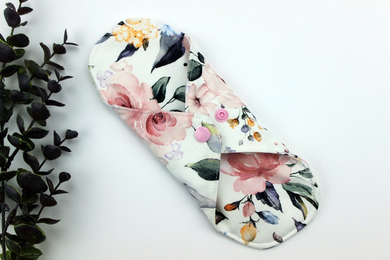 May include: A white reusable cloth menstrual pad with a floral print featuring pink, yellow, and blue watercolour flowers. The pad has two pink snaps.