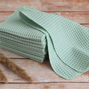 May include: A stack of light green waffle weave towels. The towels are neatly folded and stacked on a wooden surface. The towels have a textured, grid-like pattern. Dried ornamental grass is in the foreground.