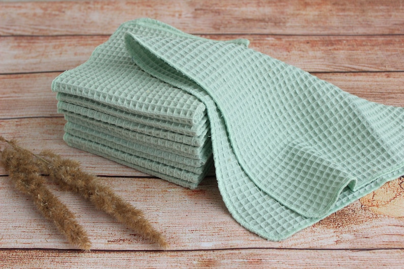 May include: A stack of light green waffle weave towels. The towels are neatly folded and stacked on a wooden surface. The towels have a textured, grid-like pattern. Dried ornamental grass is in the foreground.