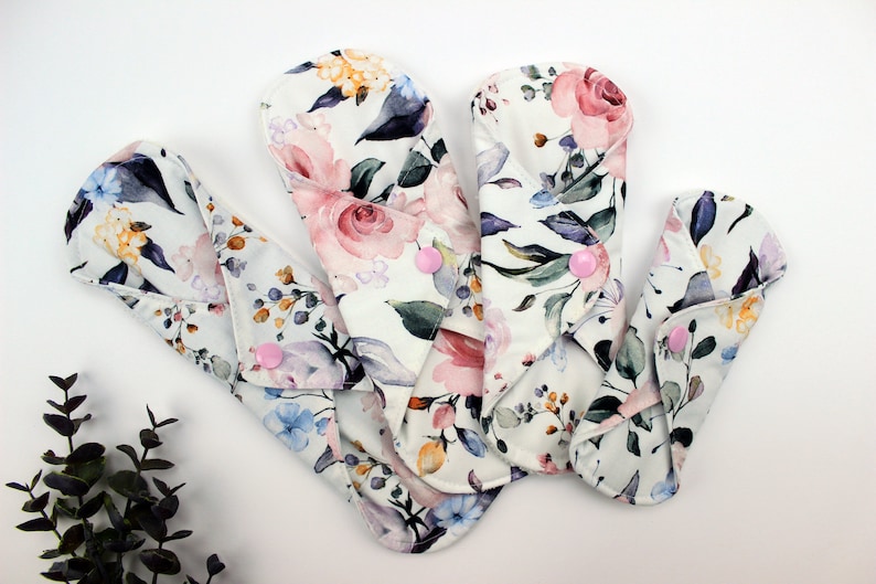 May include: Four reusable cloth menstrual pads with a floral print in shades of pink, purple, blue, and green. The pads have pink poppers.