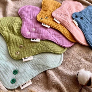 Könnte beinhalten: Sechs wiederverwendbare Stoffbinden in verschiedenen Farben, darunter Grün, Hellblau, Lila, Rosa, Gelb und Blau. Die Binden haben eine gesteppte Textur und sind in einer Reihe angeordnet. Jede Binde hat ein kleines Etikett mit dem Text "♡♡♡" darauf.