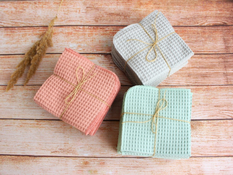 May include: Three stacks of square waffle-weave towels in pastel colors: pink, gray, and mint green. Each stack is tied with twine. The towels are arranged on a wooden surface, creating a visually appealing display of home textiles.