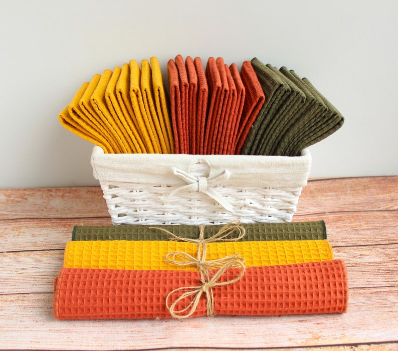 May include: A white wicker basket filled with folded waffle-weave towels in shades of yellow, orange, and olive green. Rolled towels in matching colors are tied with twine. The towels are arranged on a wooden surface.