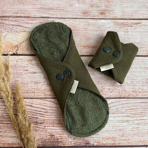 May include: Two green reusable cloth menstrual pads. The larger pad is folded in half and has two black snaps. The smaller pad is folded in thirds and has two black snaps. Both pads are made of a soft, absorbent fabric.
