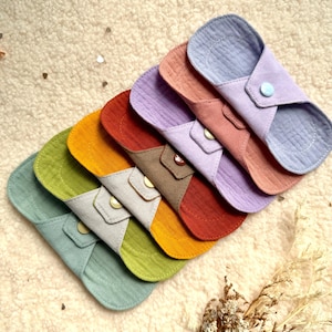 May include: A collection of reusable cloth menstrual pads in various colors, including blue, pink, purple, red, yellow, and green. Each pad features a snap closure. The pads are arranged on a textured, off-white surface.