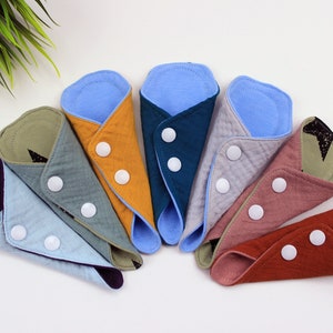 May include: A set of eight reusable cloth menstrual pads in various colors, including purple, blue, green, yellow, teal, gray, pink, and brown. Each pad has a white button closure.
