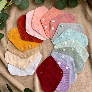 May include: A set of 12 colorful fabric bibs with snap closures. The bibs are made of a soft, textured material and come in a variety of colors, including red, orange, yellow, green, blue, purple, pink, and white.