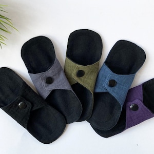 May include: Five reusable cloth menstrual pads in various colors, including black, gray, green, blue, and purple. Each pad has a black snap button.