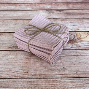 May include: A stack of light purple waffle-weave washcloths tied with twine. The washcloths are neatly folded and stacked, creating a compact bundle. The twine adds a rustic touch to the presentation. The background is a light brown wooden surface.