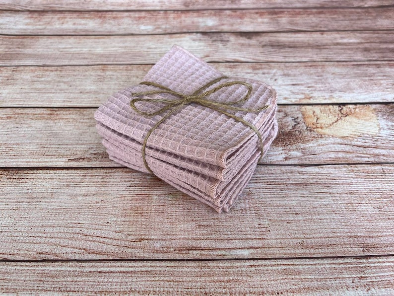 May include: A stack of light purple waffle-weave washcloths tied with twine. The washcloths are neatly folded and stacked, creating a compact bundle. The twine adds a rustic touch to the presentation. The background is a light brown wooden surface.