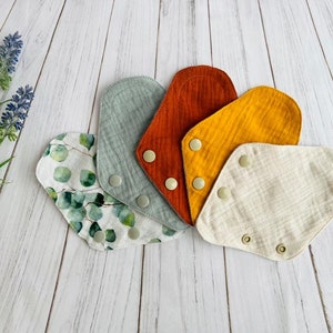May include: Five reusable cloth pads in various colors and patterns, including green with a leaf print, beige, yellow, orange, and gray. The pads have snaps on the sides.