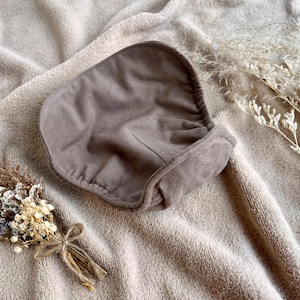 Puede incluir: Un gorro de bebé color topo con un diseño fruncido, sobre una manta suave y texturizada. También hay un pequeño ramo de flores secas atadas con hilo y una ramita de hierba ornamental seca.