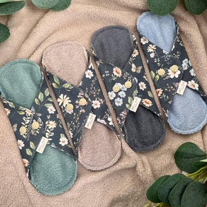 May include: Four reusable cloth menstrual pads with a floral print and different colored fabric on the back. The pads are folded in half and laid out on a brown towel.
