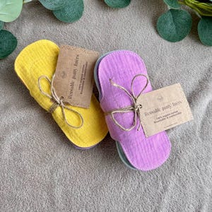 Peut inclure: Deux ensembles de protège-slips réutilisables, un ensemble est jaune et l'autre est violet. Les protège-slips sont fabriqués en coton biologique 100% et sont respirants et confortables. L'emballage est en papier brun et porte l'inscription "Protège-slips réutilisables".