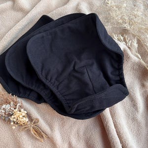 Puede incluir: Tres cubiertas de pañales de tela negras apiladas sobre una manta beige. Las cubiertas tienen una forma curva con bordes elásticos. Flores secas y espigas de trigo están dispuestas sobre la manta.