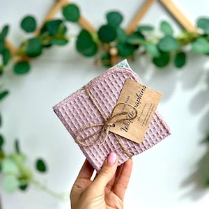 May include: A stack of pink waffle napkins tied with twine, with a small wooden tag that reads "Waffle Napkins". The napkins have a textured, square pattern. The background includes greenery and a wooden trellis.
