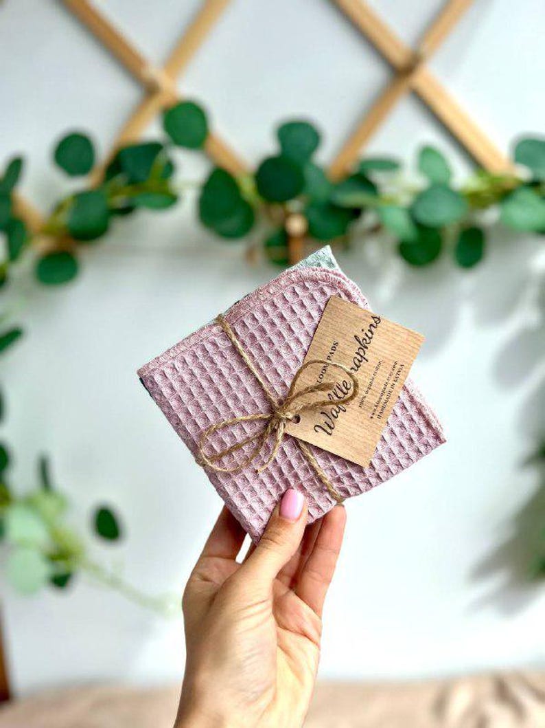 May include: A stack of pink waffle napkins tied with twine, with a small wooden tag that reads "Waffle Napkins". The napkins have a textured, square pattern. The background includes greenery and a wooden trellis.