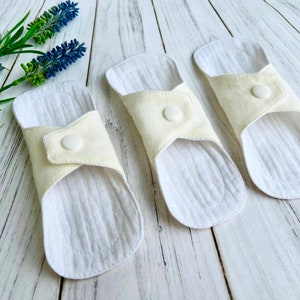 May include: Three white reusable cloth menstrual pads with a cream colored fabric flap and a white snap closure.