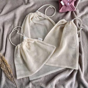 Può includere: Tre sacchetti a rete beige di diverse dimensioni con chiusure a coulisse. I sacchetti sono realizzati in un materiale leggero e traspirante e sono disposti su una superficie beige morbida e strutturata. Sullo sfondo è visibile un piccolo assorbente riutilizzabile rosa.