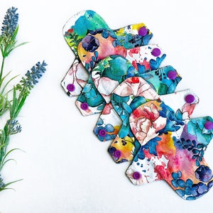 May include: A stack of five reusable cloth menstrual pads with a colorful floral print. The pads have purple snaps on the sides.