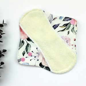 May include: A white reusable cloth pad with a floral print and a yellow microfiber insert. The pad has pink snaps on the corners.