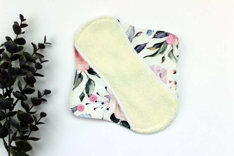 May include: A white reusable cloth pad with a floral print and a yellow microfiber insert. The pad has pink snaps on the corners.