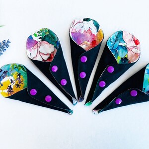 May include: Five reusable cloth menstrual pads with floral patterns. Each pad has a black outer layer with purple snaps. The pads are arranged in a fan shape.