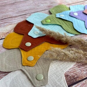 May include: A set of six reusable cloth menstrual pads in various colors, including beige, yellow, orange, red, blue, and green. The pads are layered on top of each other and have snap closures.
