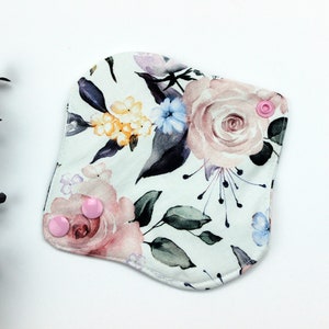 May include: A white reusable cloth pad with a floral print featuring pink, yellow, and blue flowers. The pad has two pink snap buttons.