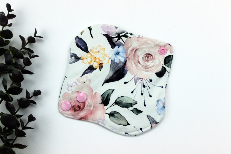 May include: A white reusable cloth pad with a floral print featuring pink, yellow, and blue flowers. The pad has two pink snap buttons.