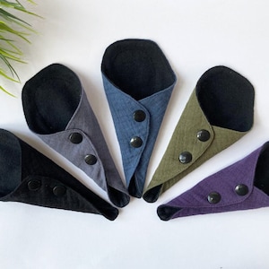May include: Five reusable cloth menstrual pads in different colors, including black, gray, blue, green, and purple. Each pad has a black button closure.