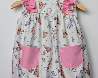 May include: White baby romper with a colorful Winnie the Pooh pattern. The sleeveless romper features pink ruffle accents on the shoulders and two pink front pockets. The romper is hanging on a wooden hanger.