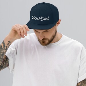 May include: A navy blue baseball cap with a white embroidered logo that says "Sad Boi".