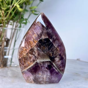 Könnte beinhalten: Ein polierter Amethyst-Kristallpunkt in Tropfenform. Der Kristall weist tiefviolette, braune und klare Quarzabschnitte auf. Der Stein wird auf einer weißen und goldenen Oberfläche mit Grün im Hintergrund ausgestellt.