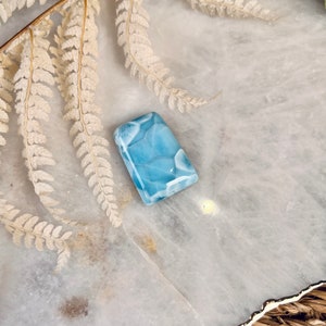 May include: A light blue, rectangular, polished, cabochon gemstone with a swirling, marbled pattern. The stone is sitting on a white, textured surface.