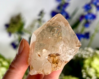 May include: A clear quartz crystal with brown inclusions. The crystal is faceted and has a pointed tip. It is being held in a hand.