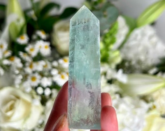 May include: A light green and blue crystal point, also known as a crystal wand, is held in a hand. The crystal is clear and has a slight shimmer to it. The background is blurred and shows white flowers.